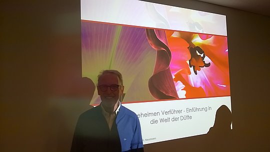 Person vor einer Projektionswand mit einer Präsentation zum Thema 'Geheimen Verführer - Einführung in die Welt der Düfte' und einem Bild einer roten Blüte.