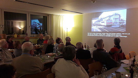 Ältere Menschen sitzen in einem dunklen Raum und schauen auf eine Wandprojektion mit einem Bild von Häusern und Text.