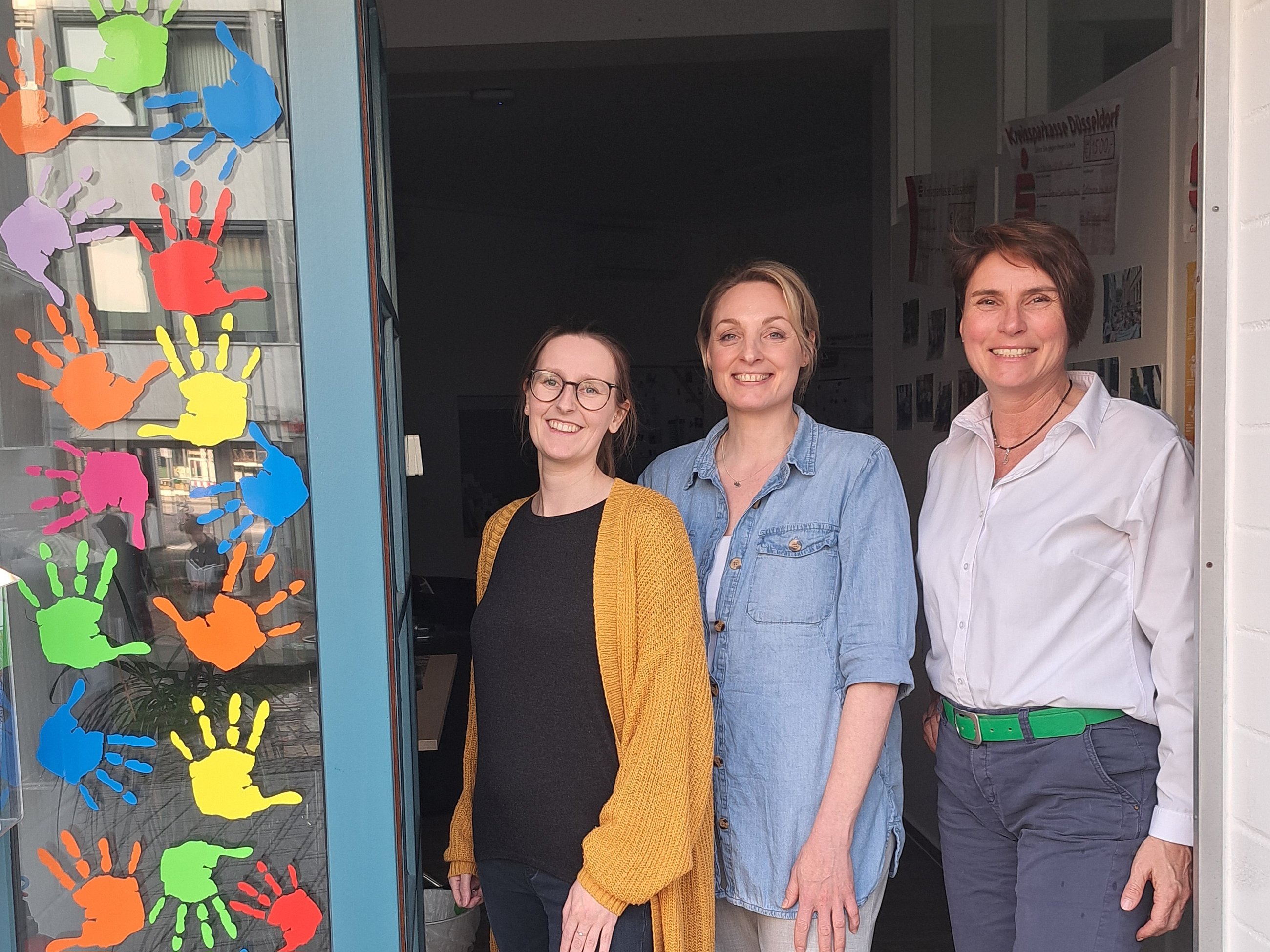 Drei Frauen stehen nebeneinander in einem Türrahmen, links bunte Handabdrücke an der Wand.