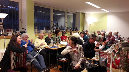 Menschen sitzen in einem Raum an Tischen, einige klatschen, eine Person spielt Akkordeon.