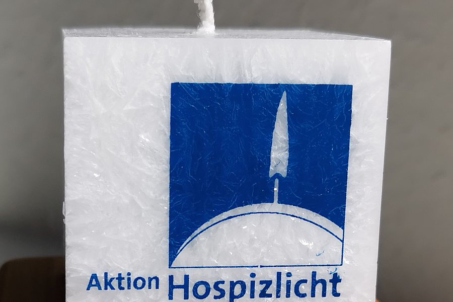 Weiße quadratische Kerze mit blauem Logo einer Flamme auf einer Halbkugel und dem Text 'Aktion Hospizlicht IN WÜRDE LEBEN – BIS ZULETZT'