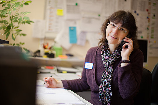 Claudia Schmitz sitzt an einem Schreibtisch, hält einen Stift in der rechten Hand und spricht am Telefon, trägt einen dunklen Pullover und einen gemusterten Schal.