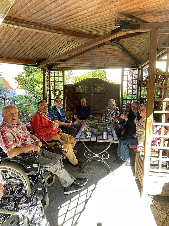 Sieben Personen sitzen in einem überdachten Gartenpavillon um einen rechteckigen Tisch mit Pflanzen darauf.