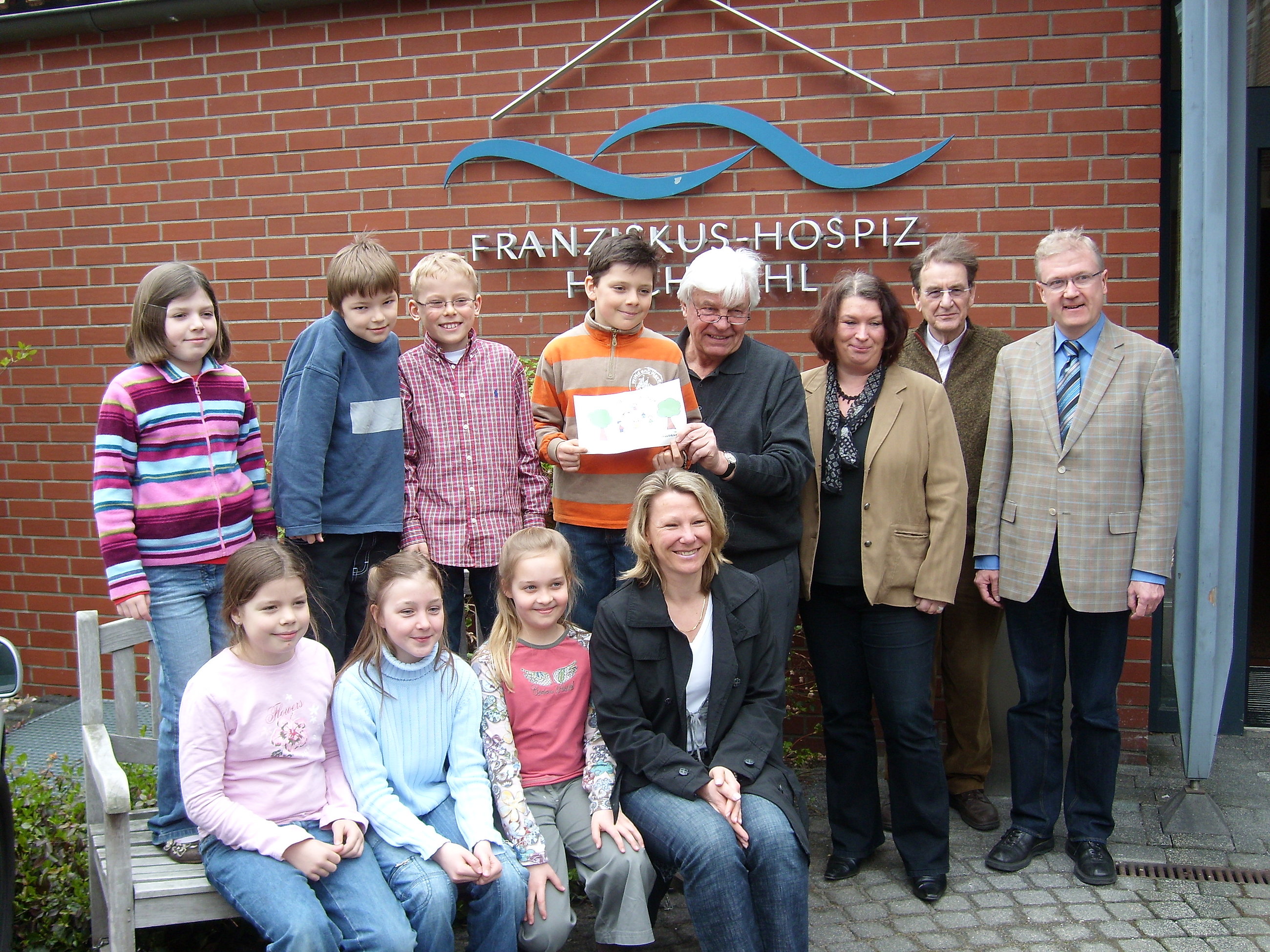Gruppe von Menschen vor roter Backsteinwand mit blauem Logo und Schriftzug 'FRANZISKUSHOSPIZ'.