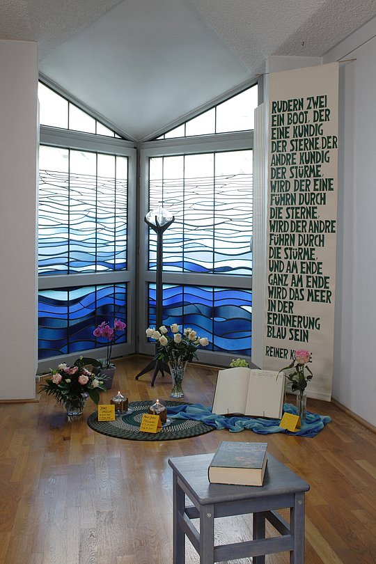 Raum mit dreiteiligem Fenster mit blauem Wellenmuster, davor Blumen, Bücher und ein aufgeschlagenes Buch auf blauem Tuch. Rechts Text auf weißem Banner.
