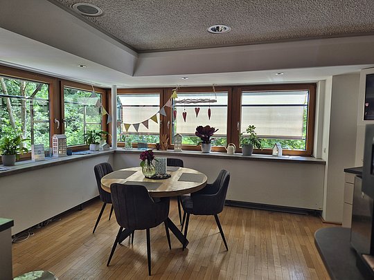 Esszimmer mit rundem Holztisch, vier gepolsterten Stühlen, Fensterbank mit Pflanzen und Dekoration, Holzboden.