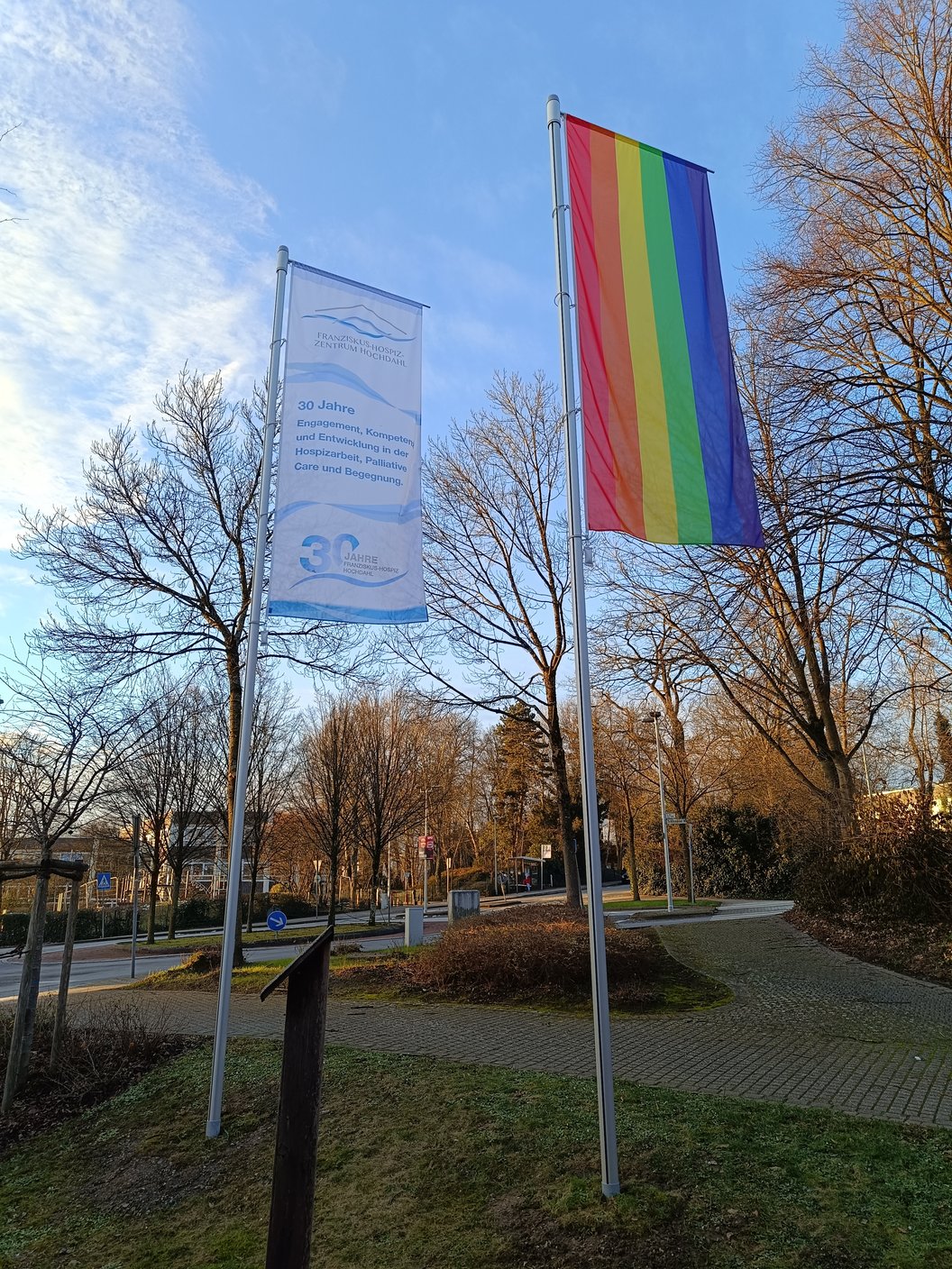 Zwei Fahnen an Masten vor kahlen Bäumen, eine mit Regenbogenfarben, die andere mit Text und Logo in Blau auf Weiß.