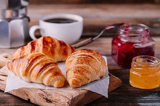 Drei Croissants auf Backpapier auf einem Holzbrett mit zwei Marmeladengläsern, einer Kaffeetasse und einem Espressokocher im Hintergrund