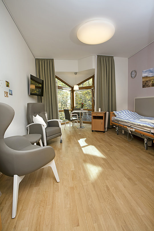 Krankenhauszimmer mit einem Bett, zwei grauen Sesseln, einem kleinen Tisch, großen Fenstern mit grünen Vorhängen und Holzboden.