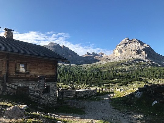 Holzhaus mit Steinfundament vor bewaldeten Bergen unter blauem Himmel