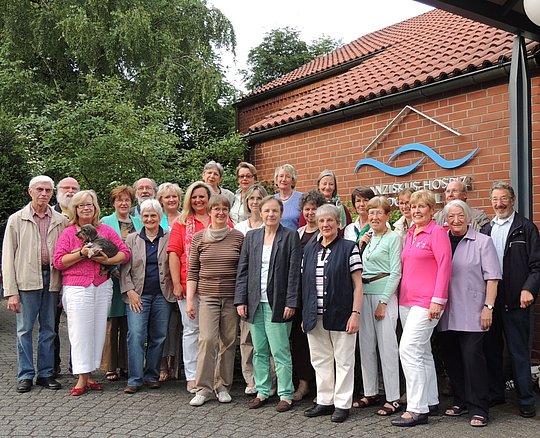 Gruppe von 20 Personen unterschiedlichen Alters steht vor einem Backsteingebäude mit rotem Ziegeldach und einem Schild mit blauem Wellenlogo.