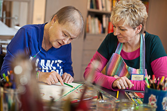 Zwei Personen sitzen an einem Tisch mit Malutensilien, eine Person hält einen grünen Stift, die andere trägt eine bunte Schürze und einen Namensschild.