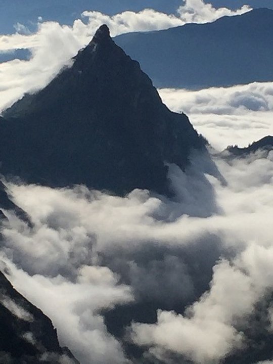 Spitze Bergspitze ragt über Wolkenmeer, dahinter weitere Bergketten unter blauem Himmel.