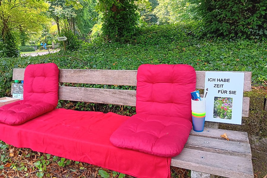 Holzbank mit roten Sitz- und Rückenpolstern, daneben ein Schild mit der Aufschrift 'ICH HABE ZEIT FÜR SIE' und bunte Becher mit Stiften