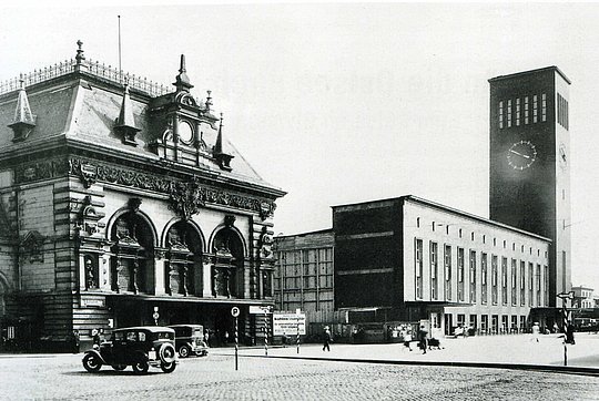 Historisches Stadtbild mit altem Gebäude im Barockstil und modernem Uhrturm, Autos und Menschen auf dem Platz.