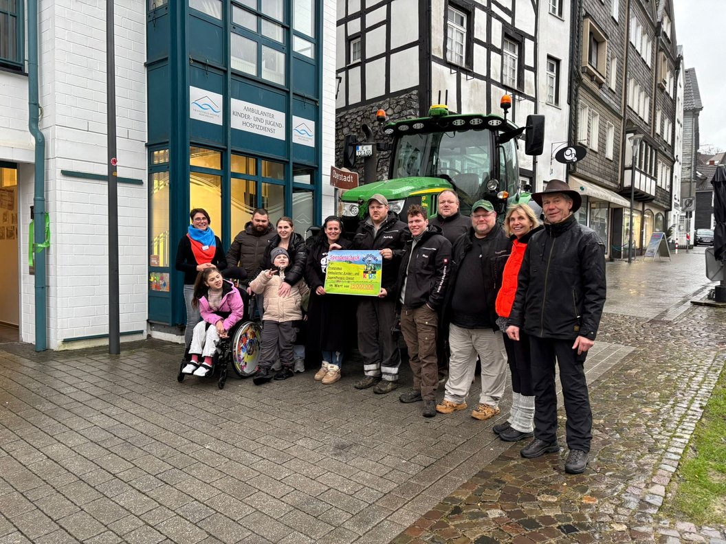 Gruppe von Menschen vor einem Fachwerkhaus und einem grünen Traktor, eine Person im Rollstuhl hält ein buntes Plakat.