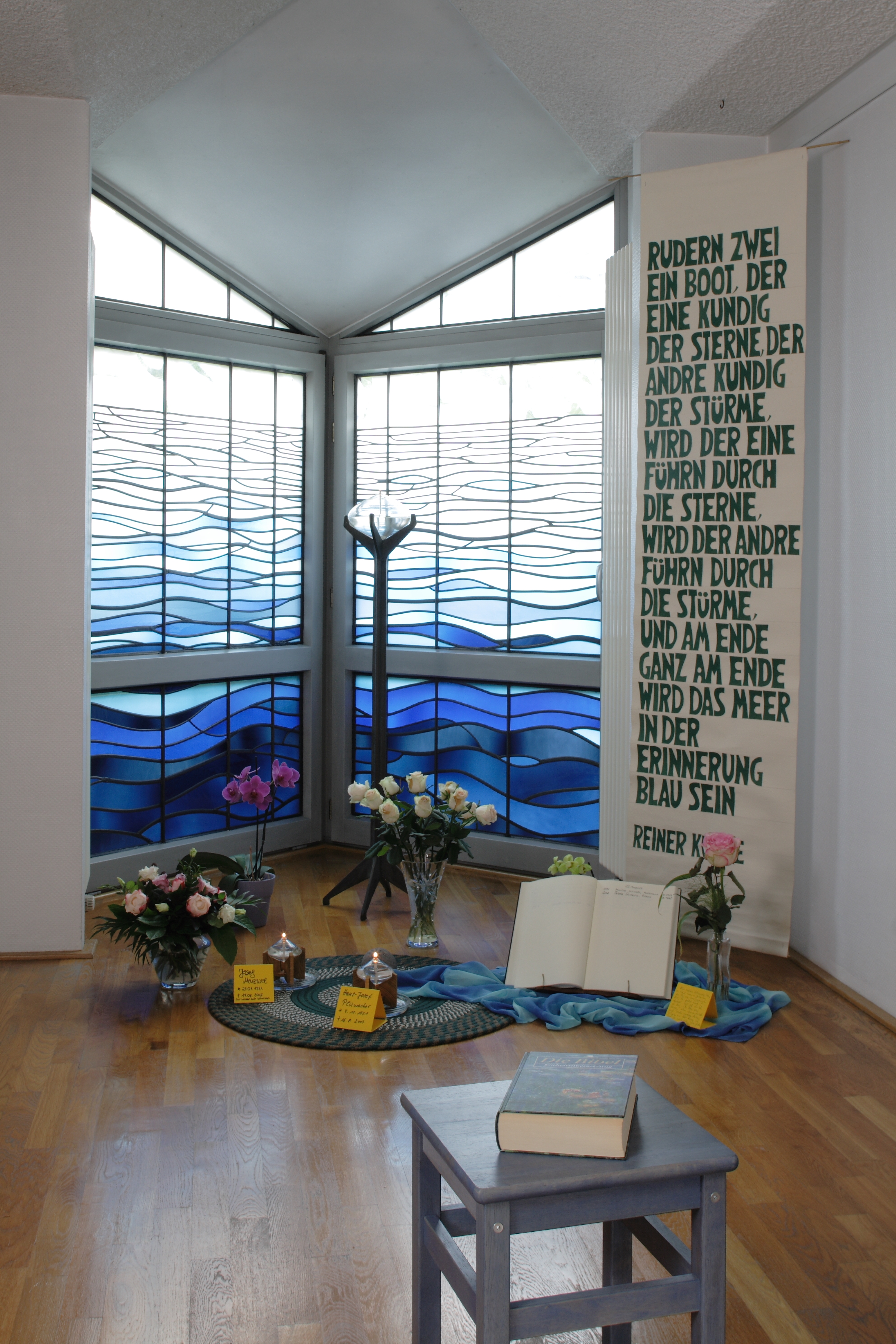 Raum mit dreiteiligem Fenster mit blauem Wellenmuster, davor Blumen, Bücher und ein aufgeschlagenes Buch auf blauem Tuch. Rechts Text auf weißem Banner.