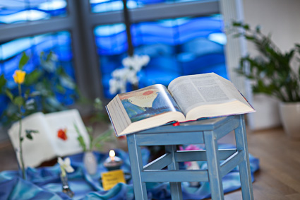 Offenes Buch auf blau gestrichenem Hocker, im Hintergrund blaue Fenster und unscharfe Pflanzen.