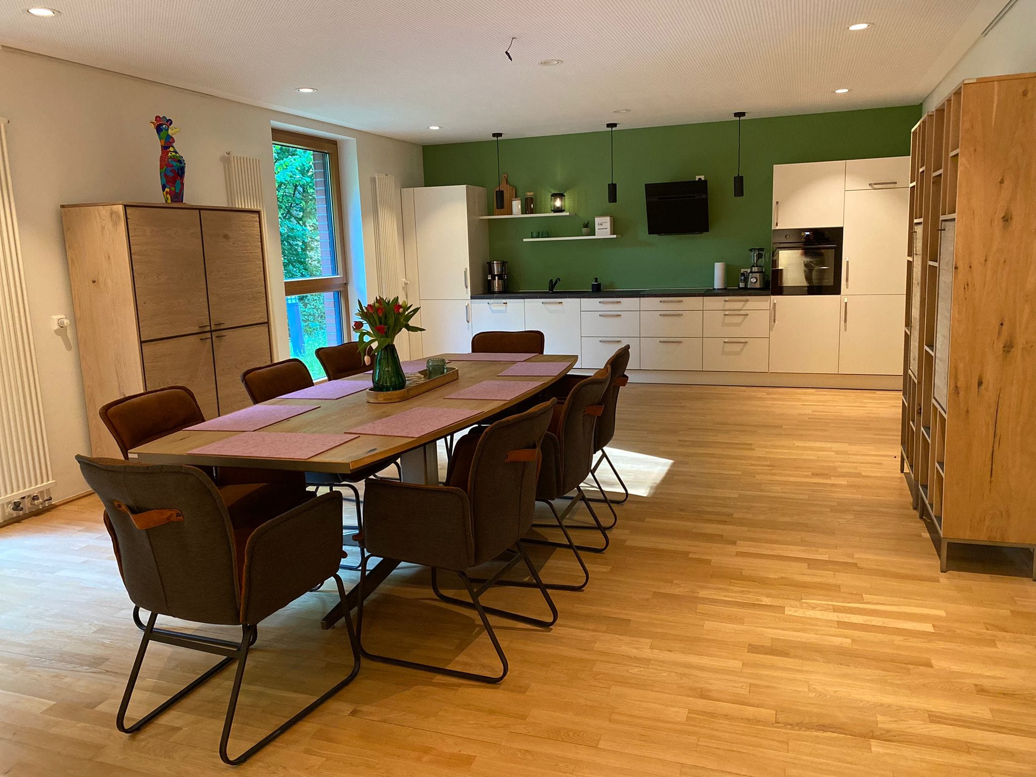 Esszimmer mit langem Holztisch, acht gepolsterten Stühlen, grüner Wand und moderner weißer Küchenzeile mit Geräten und Regalen
